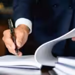 Un homme en costume noir utilisant un stylo pour signer un document sur un bureau avec une balance de justice.