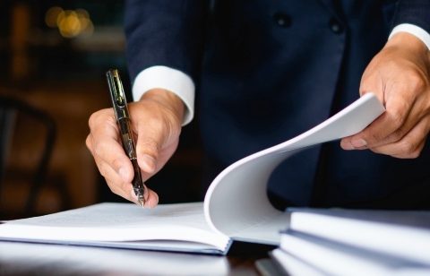 Un homme en costume noir utilisant un stylo pour signer un document sur un bureau avec une balance de justice.
