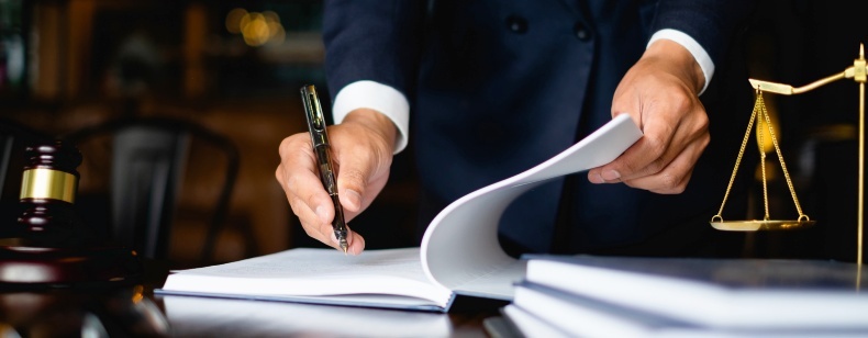 Un homme en costume noir utilisant un stylo pour signer un document sur un bureau avec une balance de justice.
