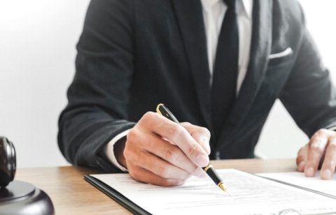 Un avocat en costume noir signant un document avec un maillet et une balance de la justice à côté.