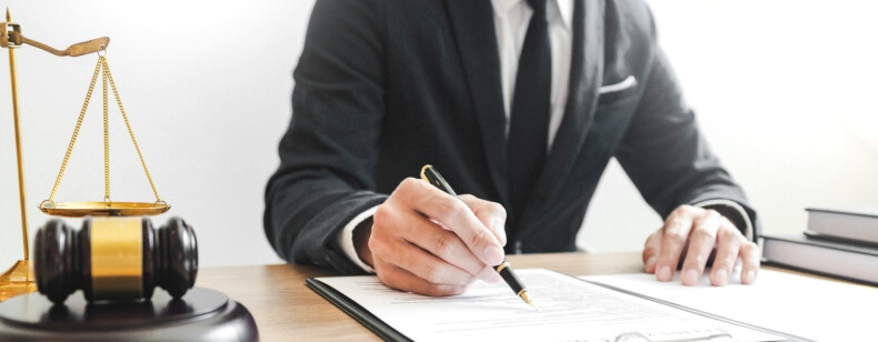 Un avocat en costume noir signant un document avec un maillet et une balance de la justice à côté.