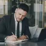Man in suit writing in notebook at table