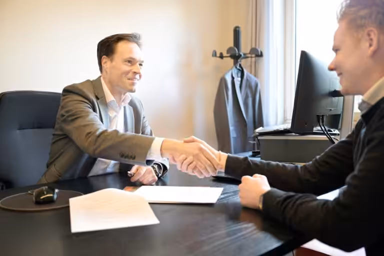 Two smiling men shake hands across an office desk